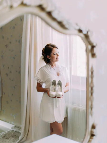 素敵な花嫁は窓を見ながら結婚式の靴を持っています。鏡に映った. — ストック写真