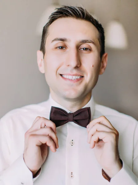Close-up portrait of the cheerful groom correcting his brown bow-tie.