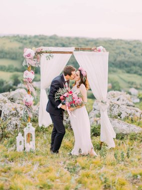 Dağlarda düğün töreni sırasında çok güzel bir şekilde dekore edilmiş arch yakınındaki öpüşme yeni evliler tam uzunlukta görünümünü.