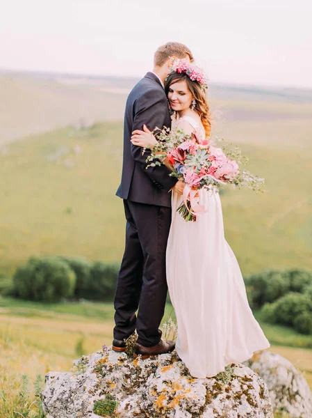 Visão completa do casal recém-casado que abraça. Groom está segurando o enorme buquê de casamento de flores coloridas . — Fotografia de Stock