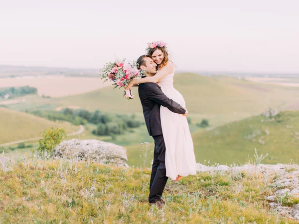 Vista horizontal do noivo sorridente carregando a noiva com o buquê de casamento nas montanhas . — Fotografia de Stock