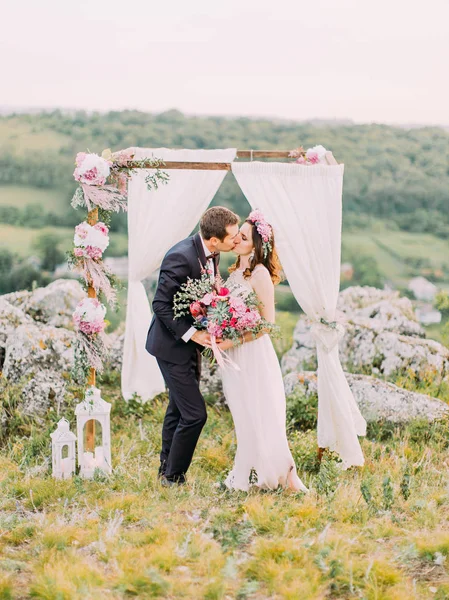 A visão completa dos recém-casados beijando perto do arco adorável decorado durante a cerimônia de casamento nas montanhas . — Fotografia de Stock
