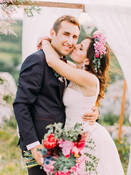 Retrato de cerca de la novia besando al novio sonriente en la mejilla durante la ceremonia de boda en las montañas . —  Fotos de Stock