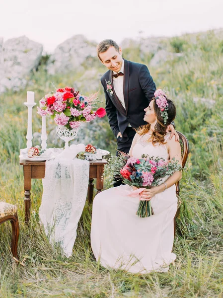 Vertical view of the bride sitting on the chair while groom is standing near her. Mountains location. — Stock Photo, Image