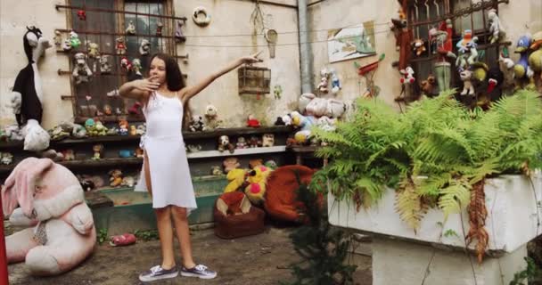 Young ballerina is stretching her body outdoor. She is raising leg up in garden with old toys and furniture. 4k footage. — Stock Video