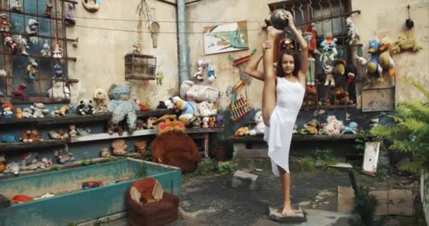 La petite fille est en train de parfumer des tours de ballet. Gymnaste mignon est debout sur une jambe avec la balle d'argent sur une autre. Des images 4k. Jardin plein de jouets et de meubles anciens emplacement . — Video