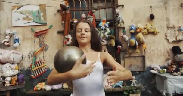 Retrato de una bonita bailarina haciendo trucos con una bola de plata en el fondo de juguetes antiguos decorando el jardín. 4k . — Vídeo de stock