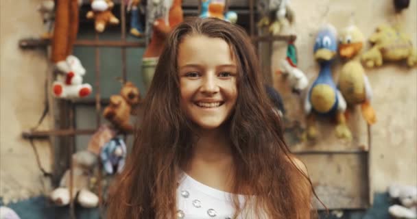 Retrato de la niña encantadora con el pelo largo mirando a la cámara y sonriendo al fondo de juguetes antiguos y vintage. Al aire libre. 4k . — Vídeo de stock