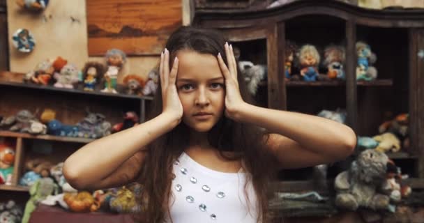 Porträt des 10-jährigen Mädchens. Ein seltsames Kind berührt ihren Kopf und blickt tief in die Kamera. Garten voller Spielzeug und alte Möbel Standort. 4k. — Stockvideo
