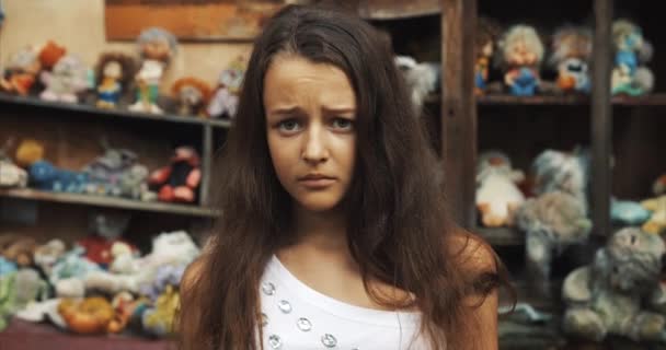 Molesto niña de 10 años de edad, poco es tristemente mirando a la cámara. Casi está llorando. jardín lleno de juguetes y muebles antiguos ubicación. 4k . — Vídeos de Stock