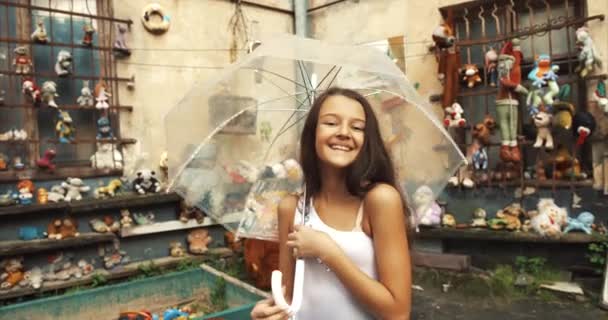 Precioso retrato de la chica bonita con linda sonrisa mirando a la cámara y girando alrededor de un paraguas transparente. Jardín lleno de juguetes antiguos ubicación. 4k . — Vídeo de stock