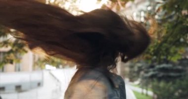 Aktif sallayarak uzun siyah saçları sevimli gülümseyen küçük kız portresi. Cityscape arka plan. 4 k görüntüleri. Güneş parlaması.