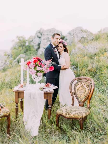 The cheerful newlywed couple is hugging behind the table set in the mountains. — Stock Photo, Image