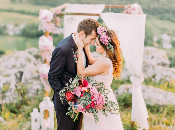 Os recém-casados abraçadores de pé nariz a nariz no fundo do arco de casamento nas montanhas . — Fotografia de Stock