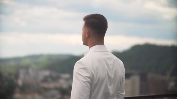 Attractive groom in white suit is coming on the terrace and enjoying the charming landscape alone. — Stock Video