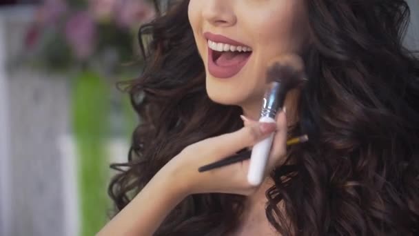 Desconocido artista de maquillaje profesional está aplicando maquillaje en la cara de la encantadora novia morena riendo con el pelo rizado . — Vídeo de stock