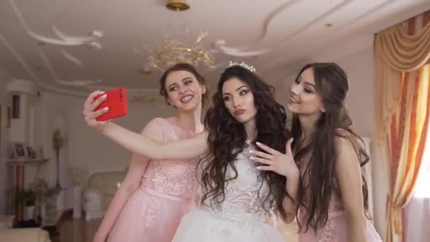 Jolie jeune mariée avec un joli maquillage et de longs cheveux bouclés prend selfies drôles avec deux belles demoiselles d'honneur à la maison . — Video