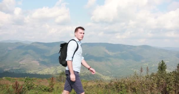 Caminar solo por las montañas. Guapo joven camina a lo largo de la colina con una mochila sobre sus hombros y mira al cielo — Vídeo de stock