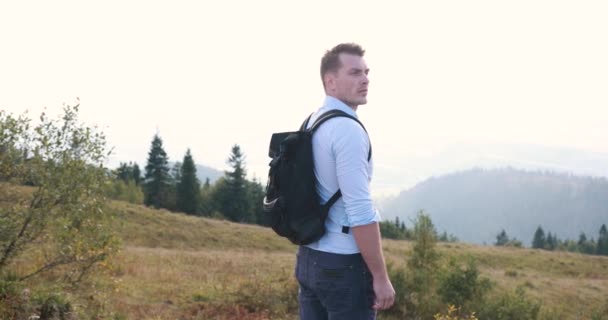 Caminar solo por las montañas. Guapo joven se para con una mochila en la cima de una colina y se ve pensativo en el paisaje de montaña — Vídeo de stock