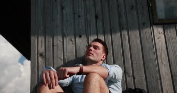 Эпическое одиночество. Красивый молодой человек сидит на крыльце старого деревянного дома на вершине холма и задумчиво смотрит в небо — стоковое видео