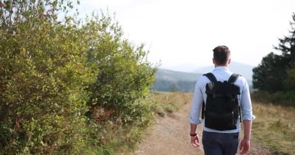 Одинокая прогулка в горах. Человек идет по дороге на холме с большим ландшафтом перед ним и смотрит через плечо — стоковое видео