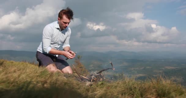 Solidão nas montanhas. Homem prepara uma fogueira no topo de uma colina com grande vista para a montanha atrás dele — Vídeo de Stock