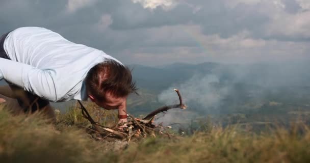 Solidão nas montanhas. Homem prepara uma fogueira no topo de uma colina com grande vista para a montanha atrás dele — Vídeo de Stock