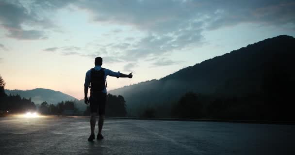 Silhueta de um homem que hitchhinking na estrada da montanha no eveninf quando os carros montam antes dele — Vídeo de Stock