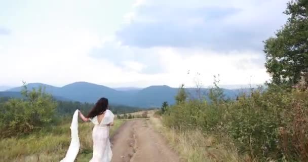 Femme en robe blanche sourit par-dessus son épaule s'enfuyant le long de la route dans les montagnes — Video