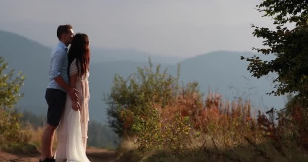 Amor nas montanhas. Homem abraça mulher por trás concurso e beija ela assistindo pôr do sol sobre as montanhas em uma colina — Vídeo de Stock