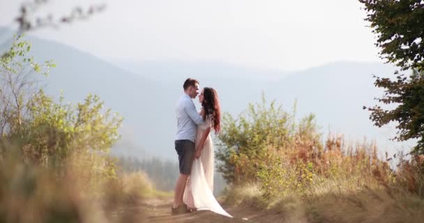 Amor en las montañas. El hombre abraza a la mujer por detrás tierna y la besa mirando el atardecer sobre las montañas en una colina — Vídeo de stock