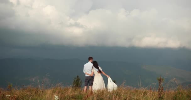 Älskar i bergen. Man och kvinna håller varandra i handen stående på backen och titta på regnet långt borta vid horisonten — Stockvideo