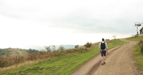 Одиночество в горах. Красивый молодой человек с рюкзаком ходит по горной дороге под тяжелым серым небом — стоковое видео