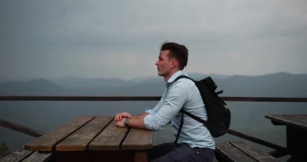 Lonely man in the mountains. Young handsome man lies his head on the arms sitting at the table with great view around him and rain falling over him — Stock Video