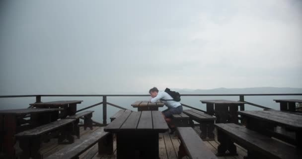 Hombre solitario en las montañas. Joven hombre guapo yace su cabeza en los brazos sentados a la mesa con una gran vista a su alrededor y la lluvia cayendo sobre él — Vídeo de stock