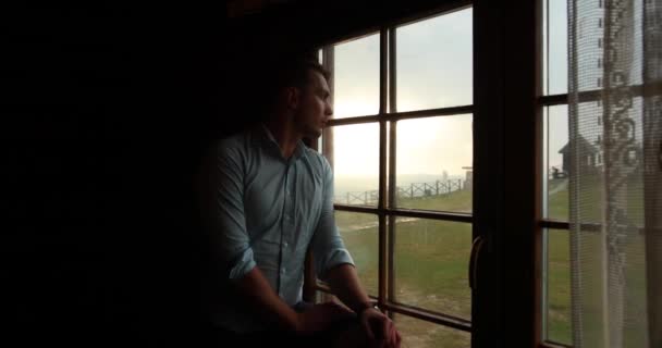 Одиночество в горах. Красивый молодой человек выглядит грустным стоя перед окном и наблюдая эпический закат над холмами — стоковое видео