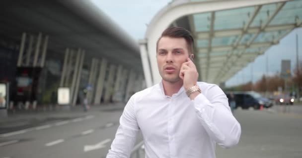 Empresario en el trabajo. Joven guapo con camisa blanca camina desde un aeropuerto con una maleta y habla por teléfono — Vídeo de stock