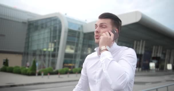 Бизнесмен на работе. Красивый молодой человек в белой рубашке выходит из аэропорта с чемоданом и разговаривает по телефону — стоковое видео
