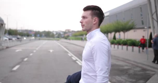 Empresário no trabalho. Bonito jovem de camisa branca caminha de um aeroporto com uma mala e olha em torno dele para um táxi — Vídeo de Stock