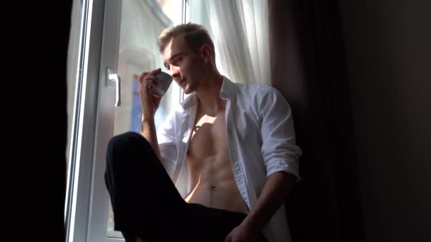 Красивий і спокусливий молодий чоловік з голим торсом п'є каву, сидячи на підвіконні вранці — стокове відео