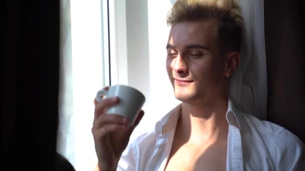 Knap en verleidelijke jongeman met naakte torso drinkt koffie zittend op de vensterbank in de ochtend — Stockvideo