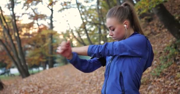Entrenando en el parque. Mujer joven con chaqueta azul se prepara para correr estirando sus brazos y piernas en el hermoso parque de otoño — Vídeo de stock