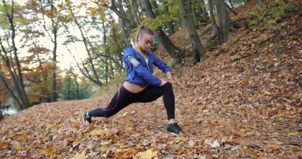 A treinar no parque. Jovem mulher de casaco azul se prepara para correr esticando os braços e pernas no belo parque de outono — Vídeo de Stock