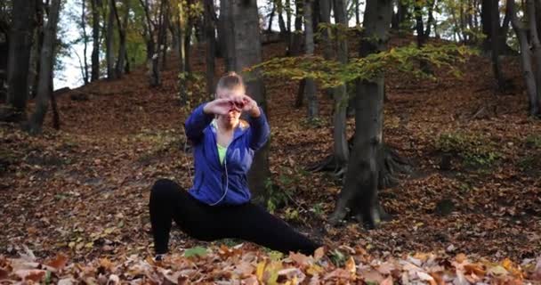 Entrenando en el parque. Mujer joven con chaqueta azul se prepara para correr estirando sus brazos y piernas en el hermoso parque de otoño — Vídeo de stock