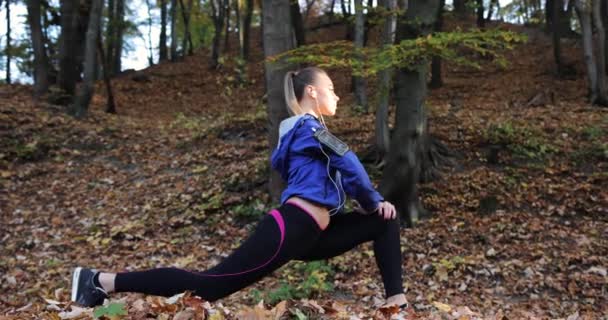 Тренируюсь в парке. Молодая женщина в синей куртке готовится к пробежке, растягивая руки и ноги в красивом осеннем парке — стоковое видео