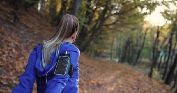 Corredor feminino no parque. Mulher bonita em casaco azul e outras roupas fitness escuta a música enquanto ela corre sobre as folhas caídas no parque cheio de sol — Vídeo de Stock