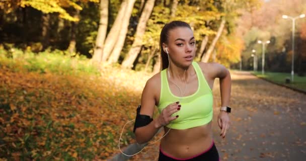 Corridore donna nel parco. Donna guarda dritto nella fotocamera mentre corre lungo il percorso nel parco autunnale coperto di foglie cadute — Video Stock