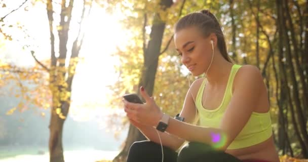 Corredor femenina en el parque. Adorable joven en traje de jogging escucha la música teniendo un descanso después de correr en un parque de otoño — Vídeos de Stock