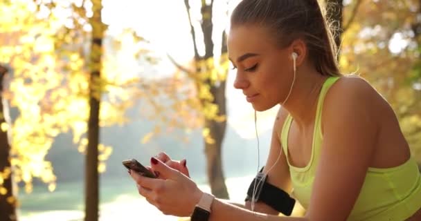 公園で女性ランナー。秋の公園で実行後休憩を持っている音楽に耳を傾けジョギング スーツで愛らしい若い女性 — ストック動画