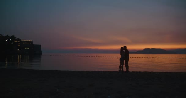 История любви на закате. Sillhouetes мужчины и женщины целуют друг друга нежно в то время как они стоят на берегу в лучах вечернего света — стоковое видео
